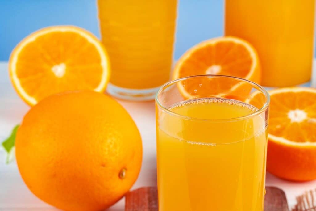 Glasses of fresh orange juice with whole and sliced oranges on a table, with a light blue background.