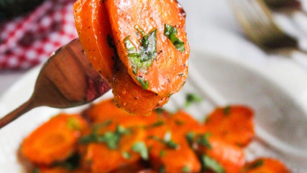 Sliced glazed carrots garnished with herbs on a fork, with more on a plate in the background.