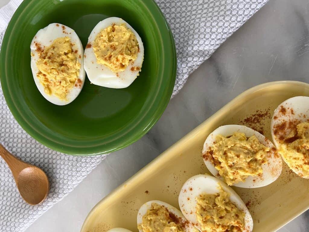 Deviled eggs served on a green plate and a yellow dish, garnished with paprika, alongside a wooden spoon on a kitchen counter.