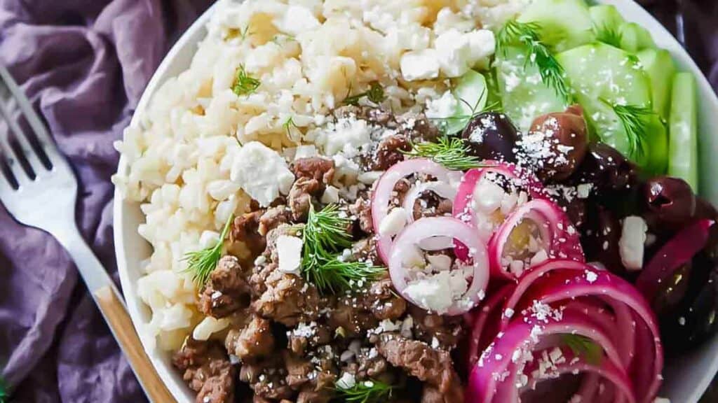 A plate of food with rice, minced meat, feta cheese, sliced cucumbers, red onions, olives, and dill, garnished with black pepper.