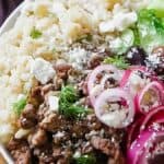 A plate of food with rice, minced meat, feta cheese, sliced cucumbers, red onions, olives, and dill, garnished with black pepper.