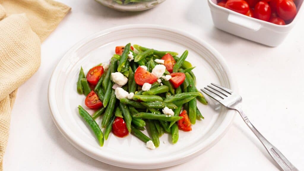 A plate of green beans tossed with cherry tomatoes and crumbled feta cheese, accompanied by a fork, on a white surface.