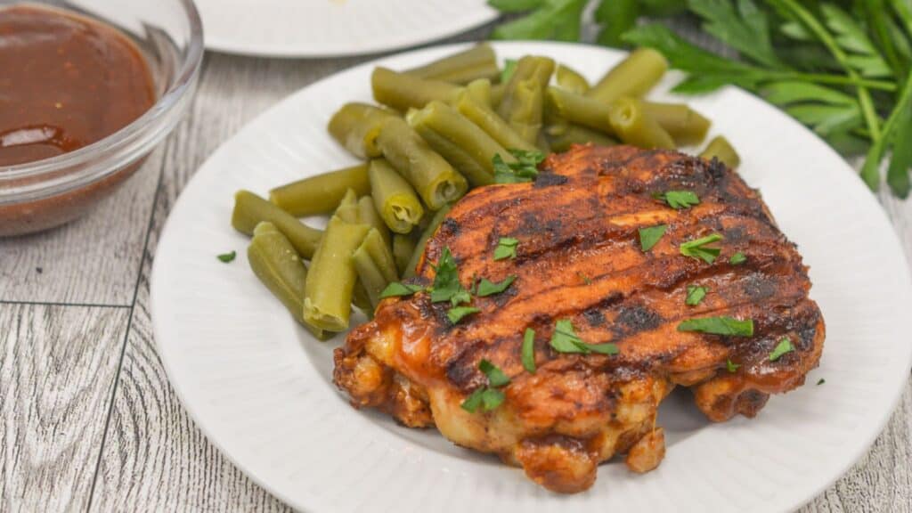 A grilled chicken thight with barbecue sauce and a side of green beans on a white plate, garnished with chopped parsley.