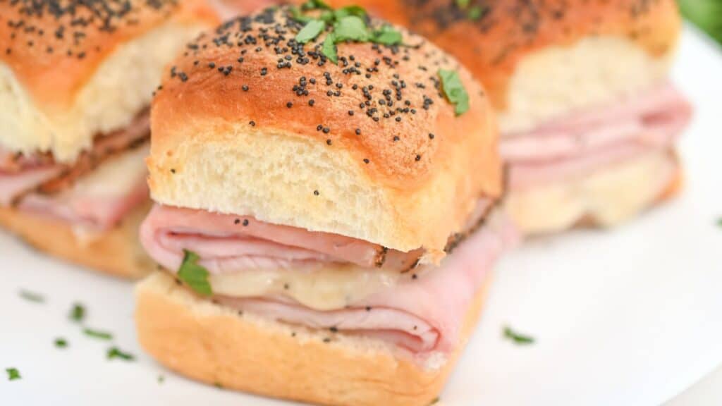 A close-up of a platter with sliced ham and cheese sliders topped with poppy seeds and sprinkled with chopped parsley.