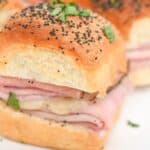 A close-up of a platter with sliced ham and cheese sliders topped with poppy seeds and sprinkled with chopped parsley.