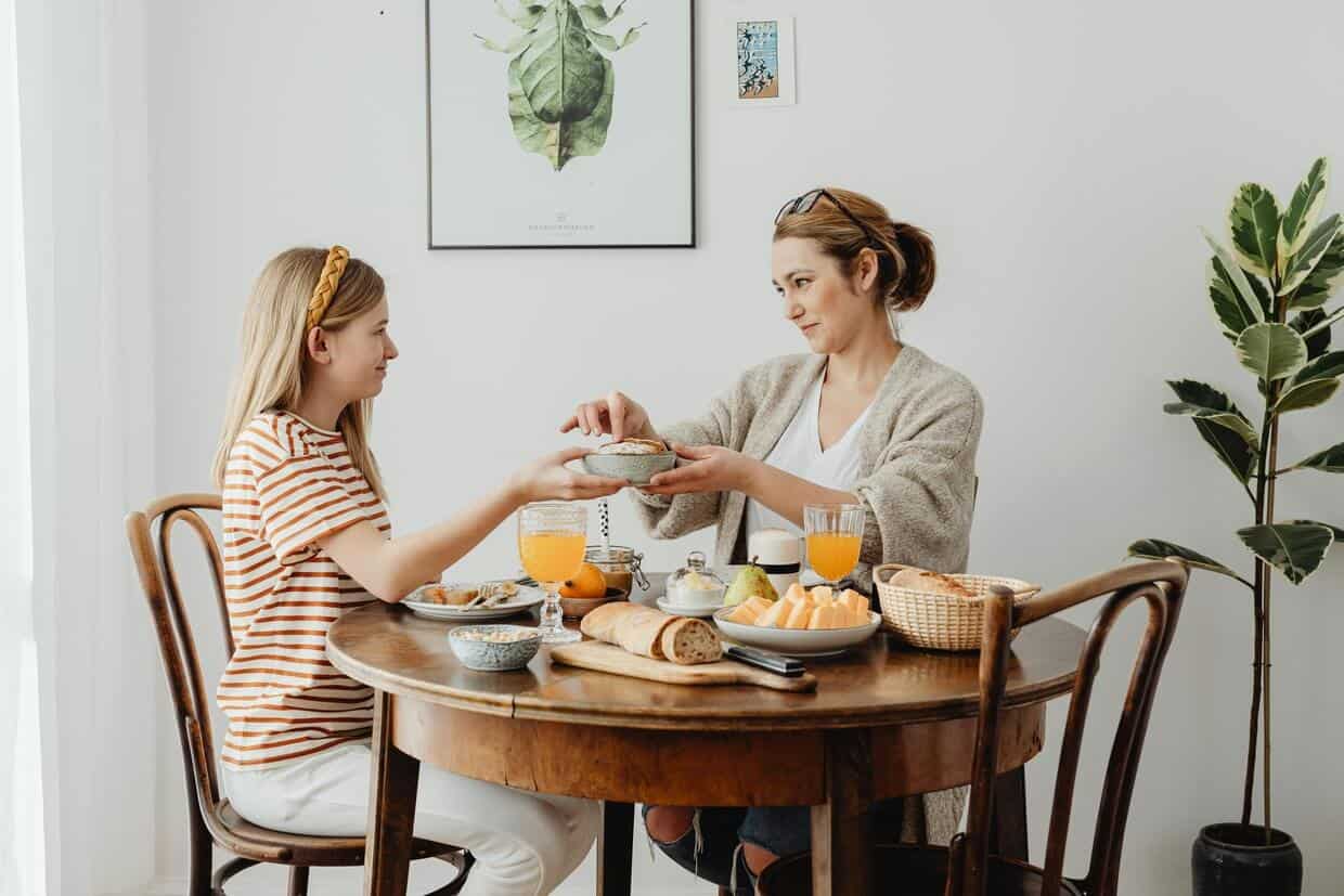 Two women sitting at a table, sharing a healthy breakfast for kids with fruit and pastries, in a light and plant-decorated room.