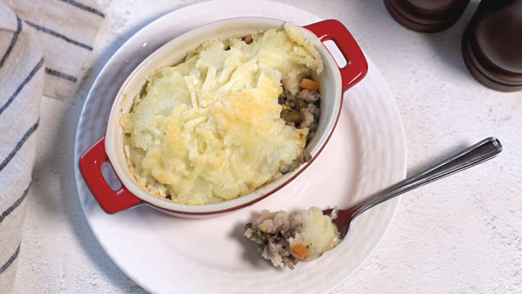A red ceramic dish of shepherd's pie with a scoop removed, showing layers of mashed potatoes and meat, on a white cloth background.