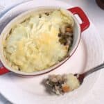 A red ceramic dish of shepherd's pie with a scoop removed, showing layers of mashed potatoes and meat, on a white cloth background.