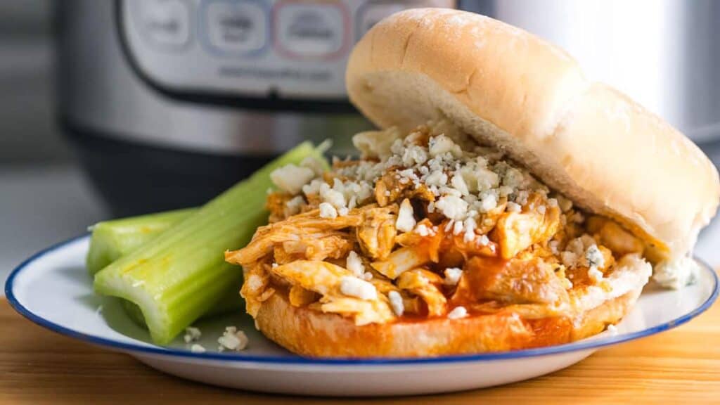 A buffalo chicken sandwich with crumbled blue cheese on a bun, served with a side of celery sticks, on a plate near an instant pot.