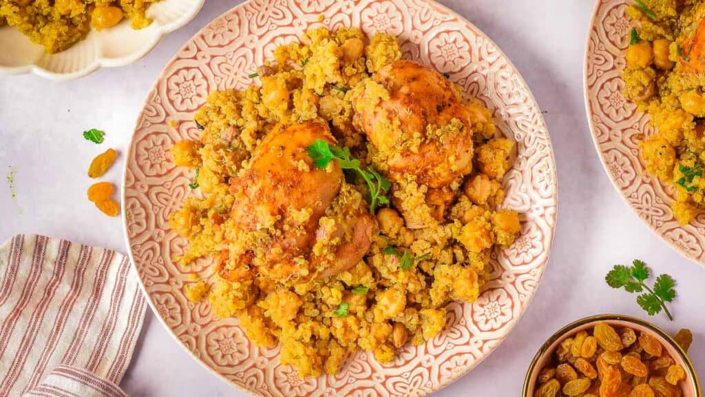 A plate of chicken thighs served over a bed of seasoned couscous with chickpeas and garnished with fresh parsley, surrounded by similar dishes and a cloth napkin.
