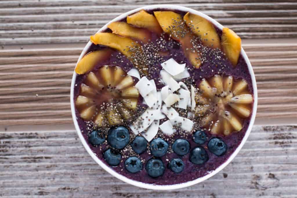 A bowl of acai smoothie topped with slices of kiwi, mango, coconut, blueberries, and chia seeds, arranged neatly on a wooden surface.