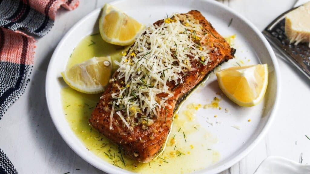 A grilled salmon fillet topped with herbs and parmesan cheese, served with lemon wedges on a white plate.