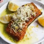 A grilled salmon fillet topped with herbs and parmesan cheese, served with lemon wedges on a white plate.