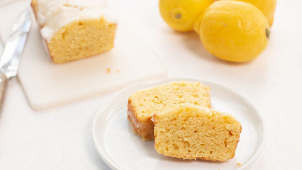 Slices of lemon loaf with icing on a white plate, with whole lemons in the background.