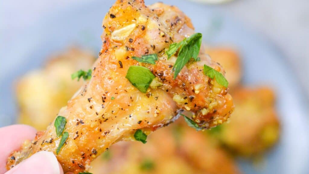 Close-up of a hand holding a seasoned chicken wing garnished with fresh herbs, with more pieces on a plate in the background.