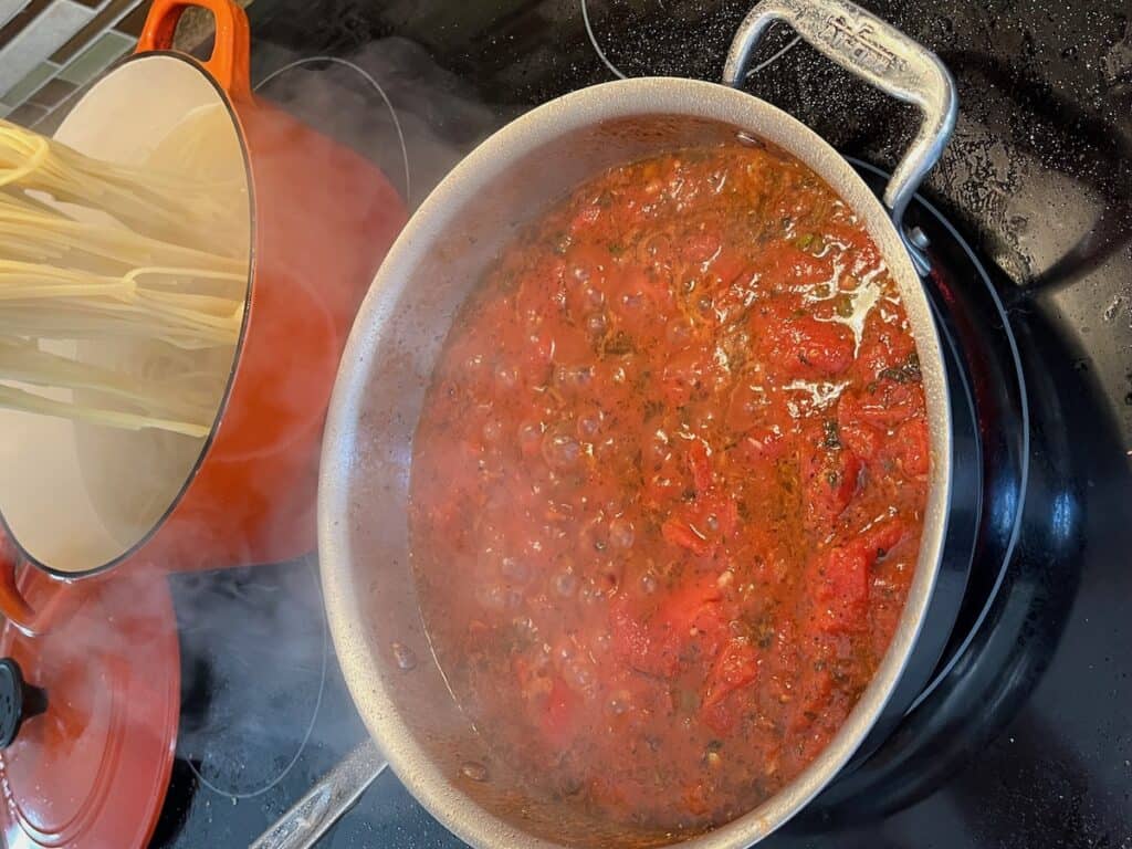 A pot of simmering tomato sauce next to another pot with boiling pasta on a stovetop, steam visible.