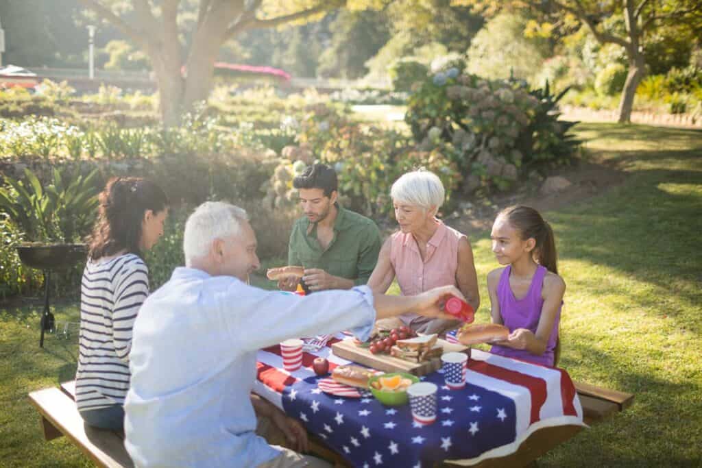 A group of adults enjoys a picnic in a sunny park, sharing food at a table decorated with a u.s. flag-themed tablecloth.