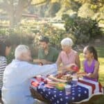 A group of adults enjoys a picnic in a sunny park, sharing food at a table decorated with a u.s. flag-themed tablecloth.