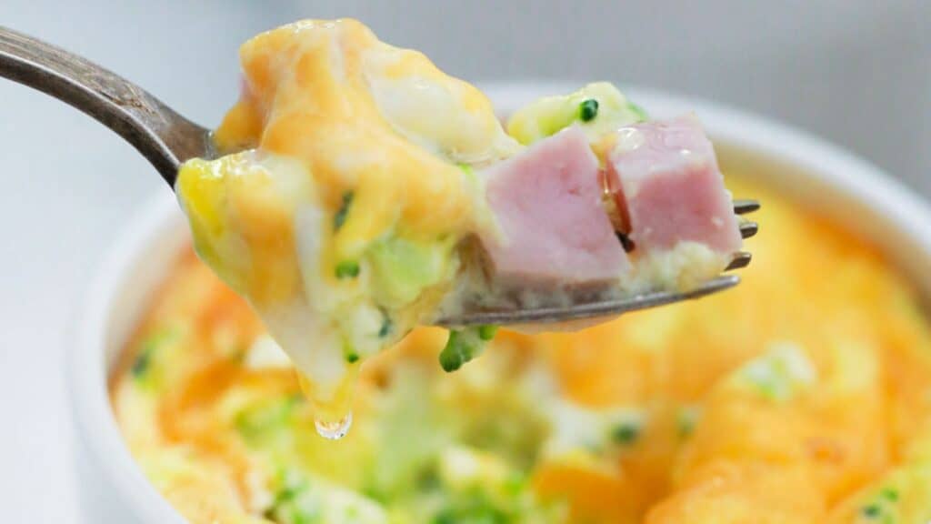 A fork holding a bite of cheesy broccoli and ham frittata with a drip of cheese sauce.