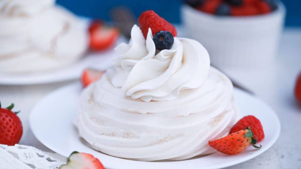 A pavlova dessert topped with whipped cream and fresh berries on a white plate, surrounded by whole strawberries.