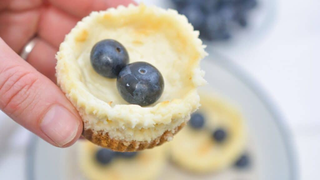Close-up of a hand holding a mini cheesecake topped with two blueberries, with more cheesecakes and blueberries on a plate in the background.