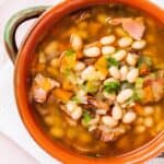 A bowl of bean soup with chunks of ham and diced vegetables, served in a rustic terracotta bowl with a spoon on the side.