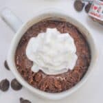 A mug cake topped with whipped cream, served beside a small jar of nutella and scattered chocolate chips on a white surface.