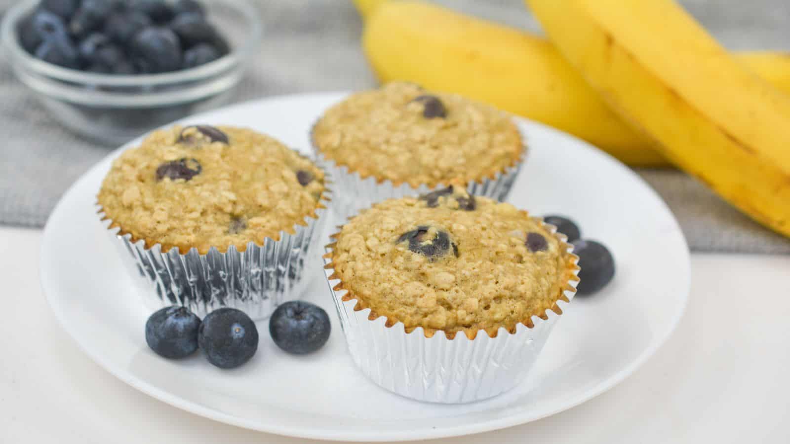 Three blueberry banana muffins on a white plate, with fresh blueberries and bananas in the background.
