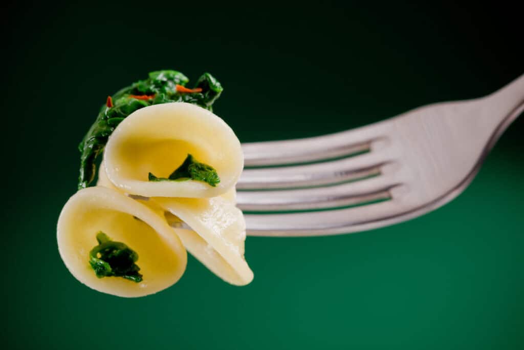 Tortellini pasta noodles with spinach on a fork, close-up, against a dark green background.