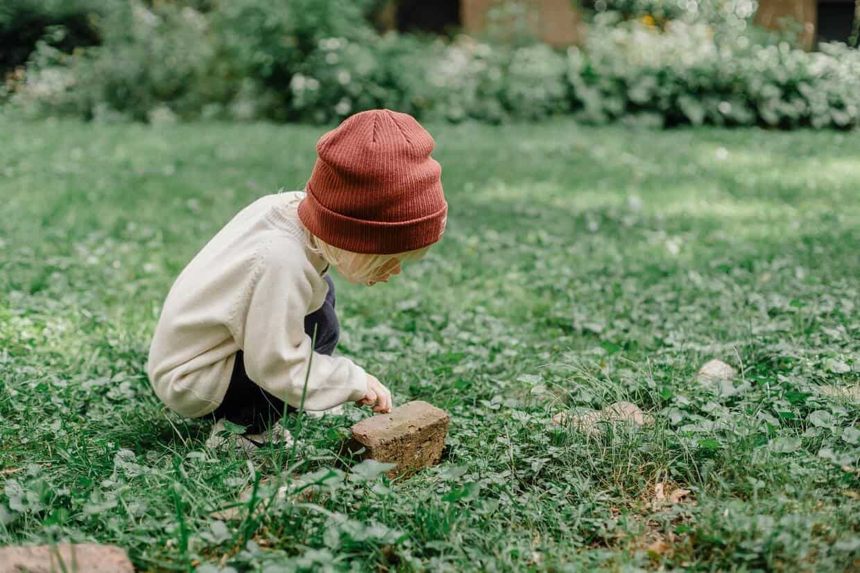 A child in a red beanie and cream sweater crouching on a grassy field, engaging in activities for preschoolers by inspecting a rock.