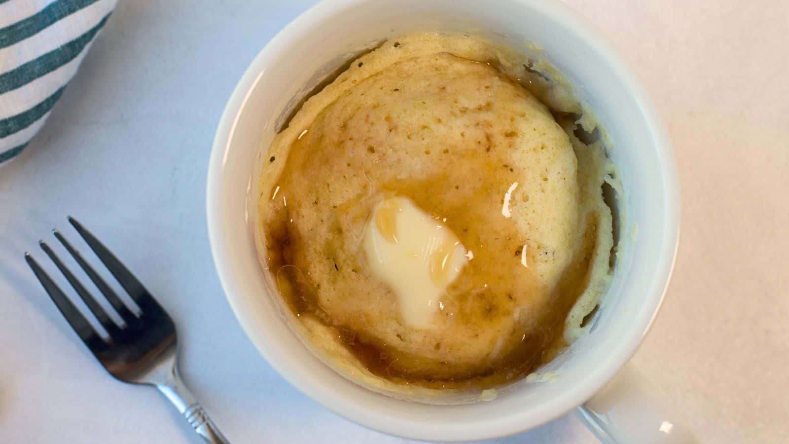 A pancake mug cake with syrup on top, viewed from above, next to a fork on a marble surface.