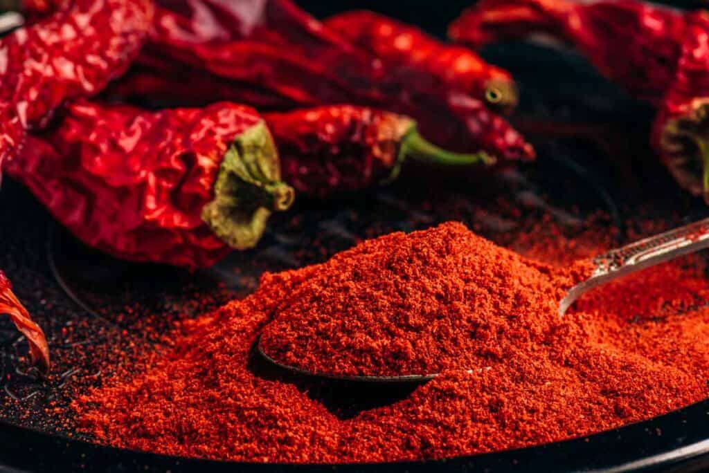 A close-up of vibrant red paprika with a spoon, surrounded by dried chili peppers on a dark background.