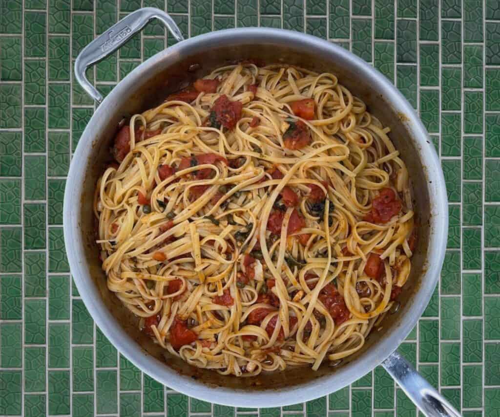 A pot of spaghetti with tomato chunks on a green tiled surface.