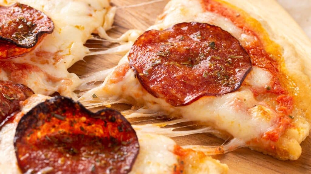 A close-up of a slice of pepperoni pizza with melted cheese and visible herbs on a wooden surface.