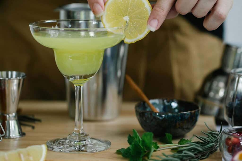 A hand squeezing a lemon over a green cocktail in a martini glass, with cocktail-making ingredients and tools in the background.