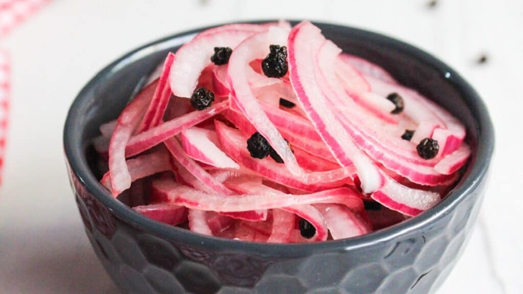 A bowl of pickled red onions garnished with black peppercorns, set on a white surface.