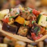 A wooden spoon holds a colorful mixture of diced vegetables including eggplant, zucchini, and peppers, topped with chopped herbs.