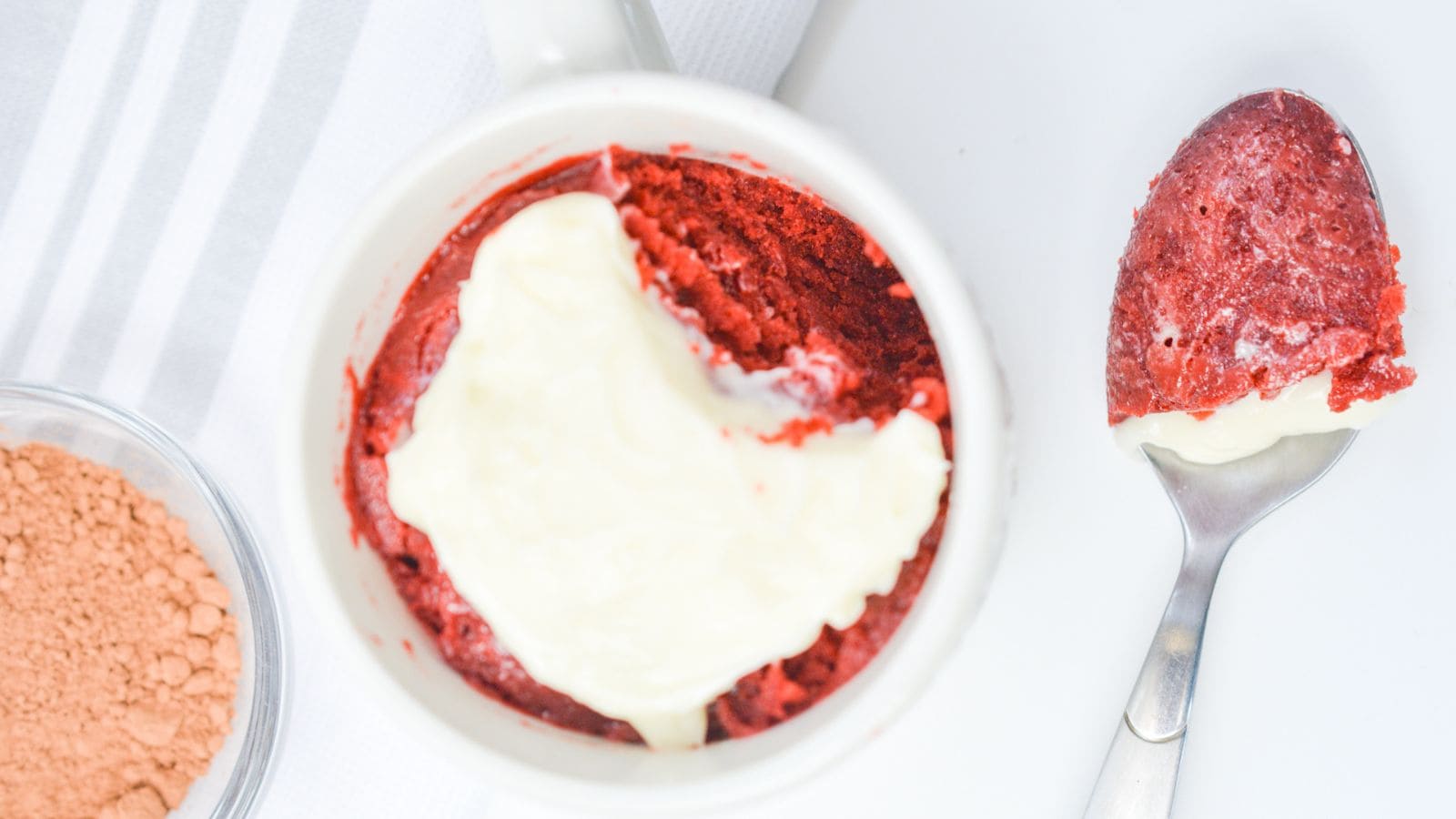 Red velvet cake with a layer of cream cheese frosting in a white ramekin, next to a spoon with a bite taken out, on a white background.
