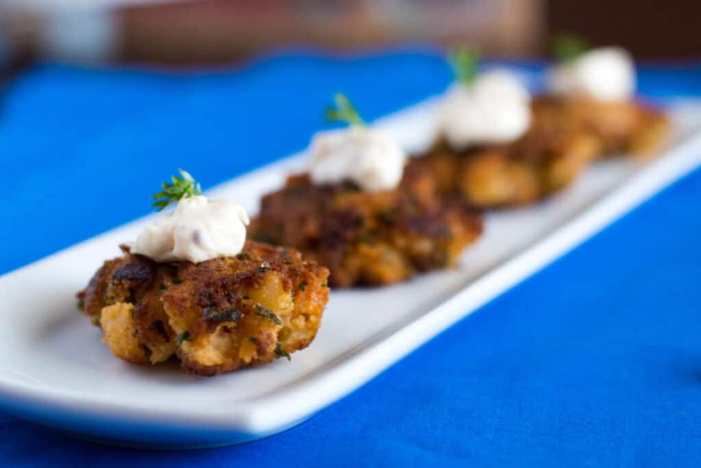 A row of crab cakes topped with a dollop of sauce on a white rectangular serving plate, set against a soft-focus blue background.