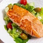 Grilled salmon fillet on a bed of lettuce with cherry tomatoes, cucumbers, and olives, served on a white plate with a fork on the side.