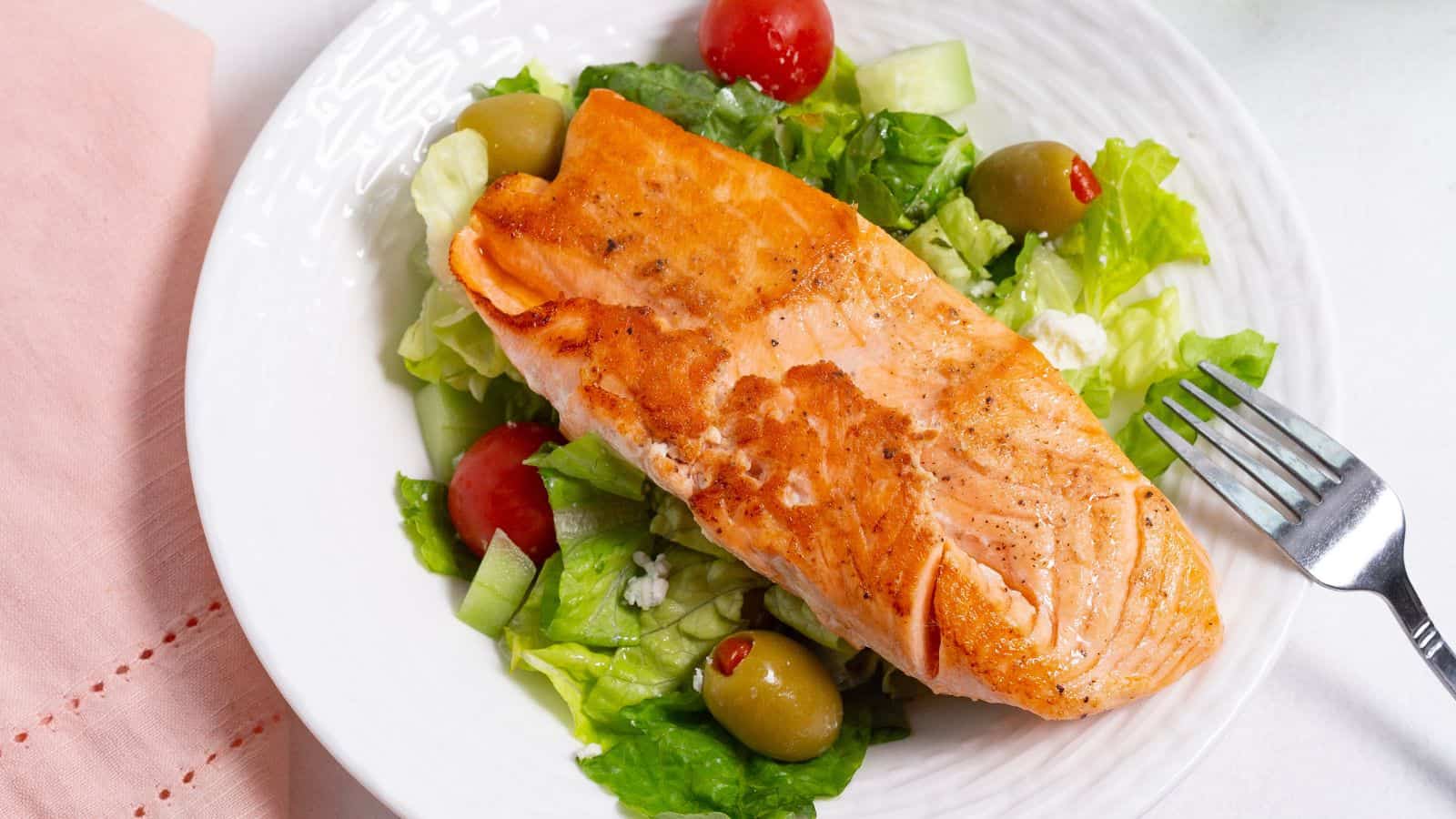 A grilled salmon fillet served on a bed of fresh salad with lettuce, tomatoes, and olives on a white plate.
