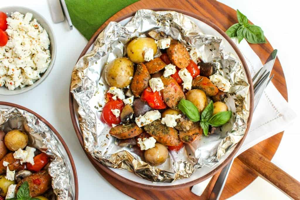 An opened foil packet on a plate with sliced chicken sausage, potatoes, vegetables, and crumbled feta cheese.