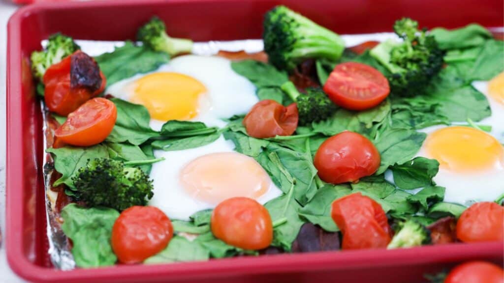 Baked eggs with spinach, broccoli, and cherry tomatoes in a red baking dish.