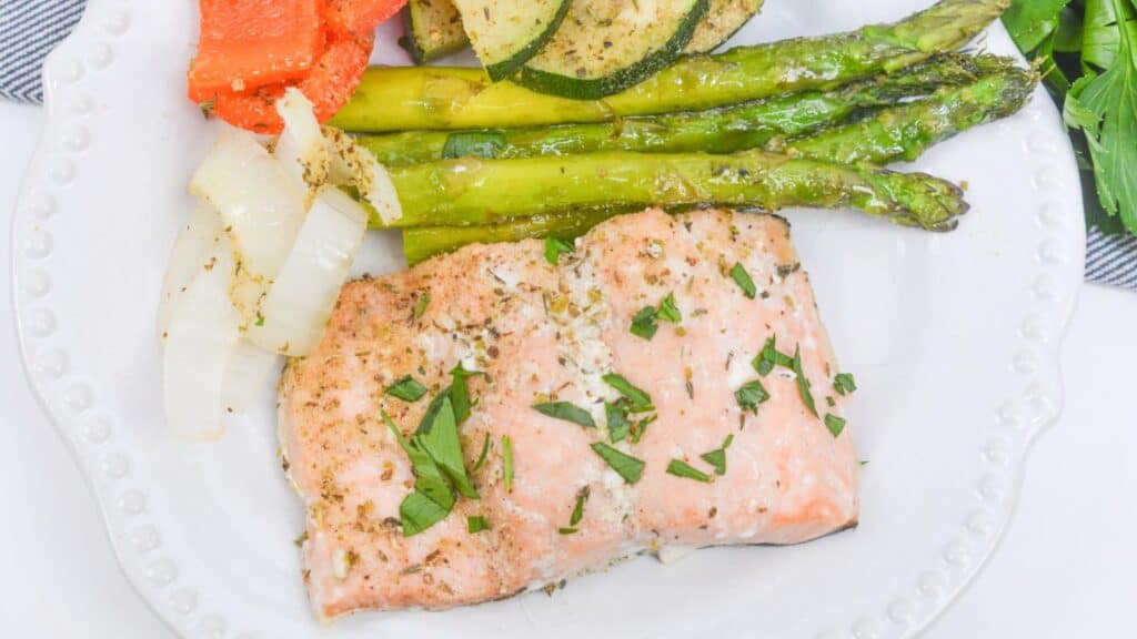 Baked salmon fillet garnished with herbs, served with asparagus, zucchini, peppers, and onions on a white plate.