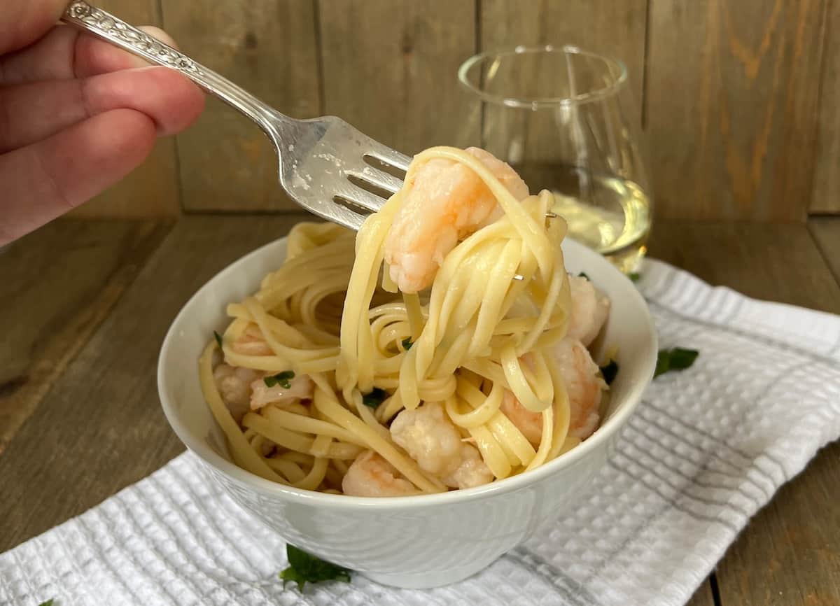A hand holding a fork twirls spaghetti with shrimp in a white bowl, with a glass of white wine in the background on a wooden table.