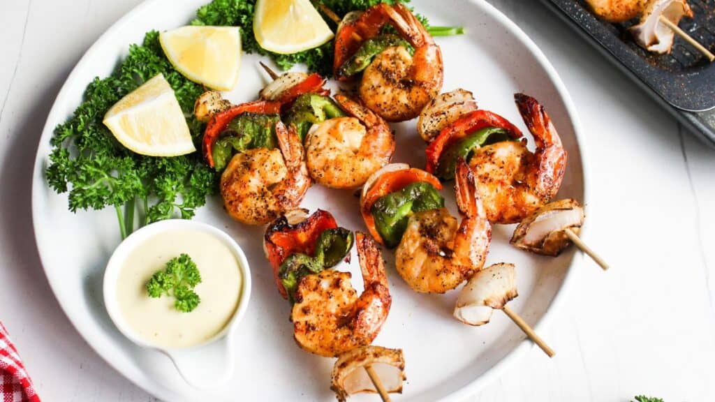Grilled shrimp skewers with bell peppers and onions, served with lemon wedges and dipping sauce on a white plate.