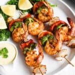 Grilled shrimp skewers with bell peppers and onions, served with lemon wedges and dipping sauce on a white plate.