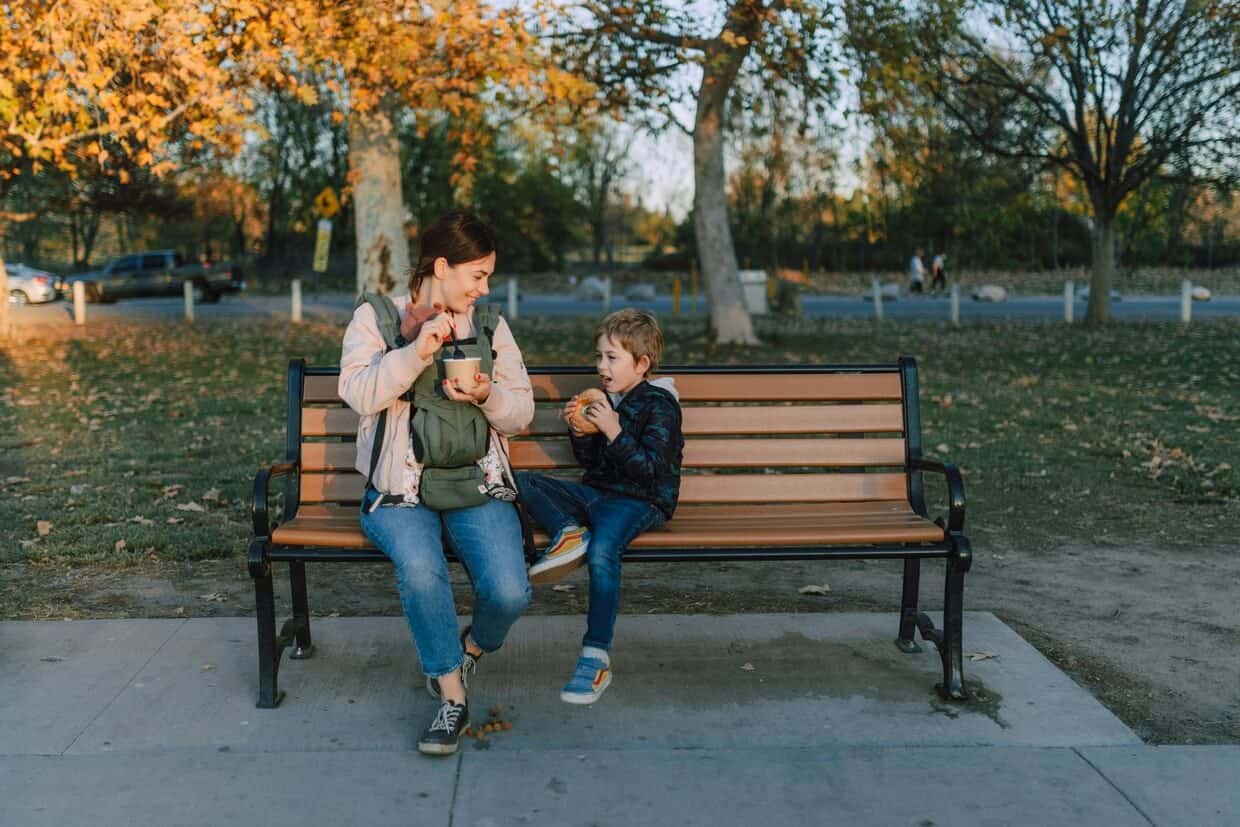A mother and her two children sitting on a park bench in autumn; she is feeding breakfast to her baby while talking to her young son.