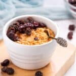 A bowl of baked oatmeal topped with raisins on a wooden table, accompanied by a blue cloth and spoon.