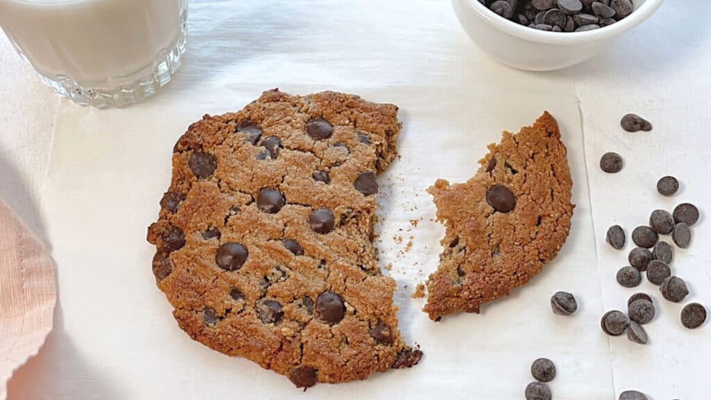 A large chocolate chip cookie broken in two pieces, next to a bowl of chocolate chips and a glass of milk on a white surface.
